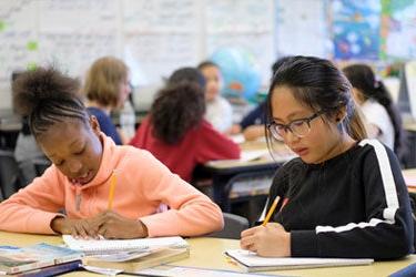 Two students work side by side at a desk in a classroom